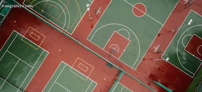 Aerial view of outdoor sports courts displaying multiple basketball and tennis courts positioned side by side. The courts are painted with green playing surfaces and red surrounding areas, with white and yellow boundary lines. The basketball courts feature hoops and painted three-point arcs and key areas, while the tennis courts include service lines and baselines.