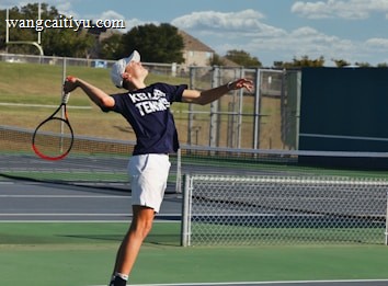 A tennis player is captured mid-air while serving at an outdoor tennis court. The player is wearing a navy blue shirt with 'Keller Tennis' printed on it, white shorts, and bright green sports shoes. A tennis racket is in the player's right hand, while the tennis ball is in the air above. The background includes a chain-link fence, a field, trees, and a partly cloudy sky.