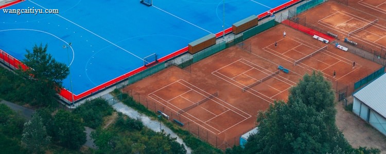 An aerial view of a large sports complex with a vibrant blue field surrounded by a red boundary and adjacent clay courts. The complex is nestled amidst dense greenery and trees, with a few buildings visible in the background. The setting appears to be serene and well-maintained, with a sense of spaciousness.